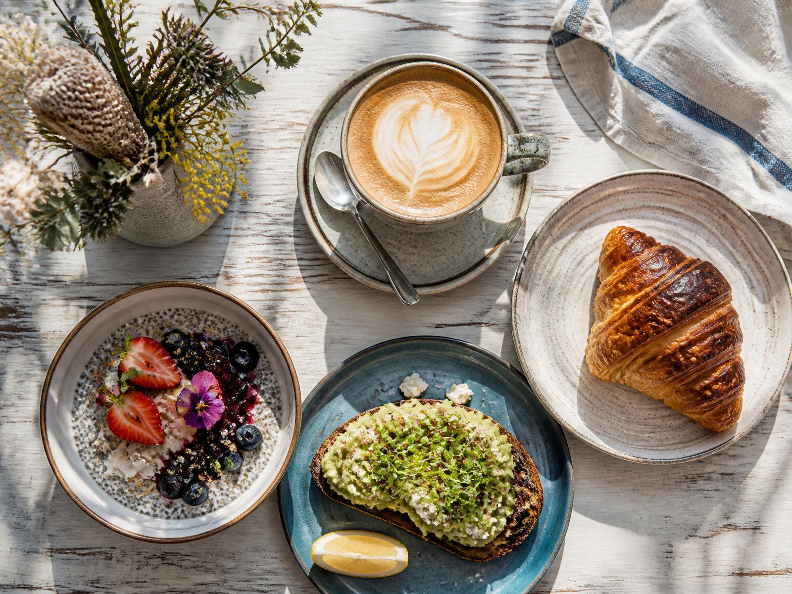 Latte, croissant, chia bowl and avocado toast on a weathered timber table